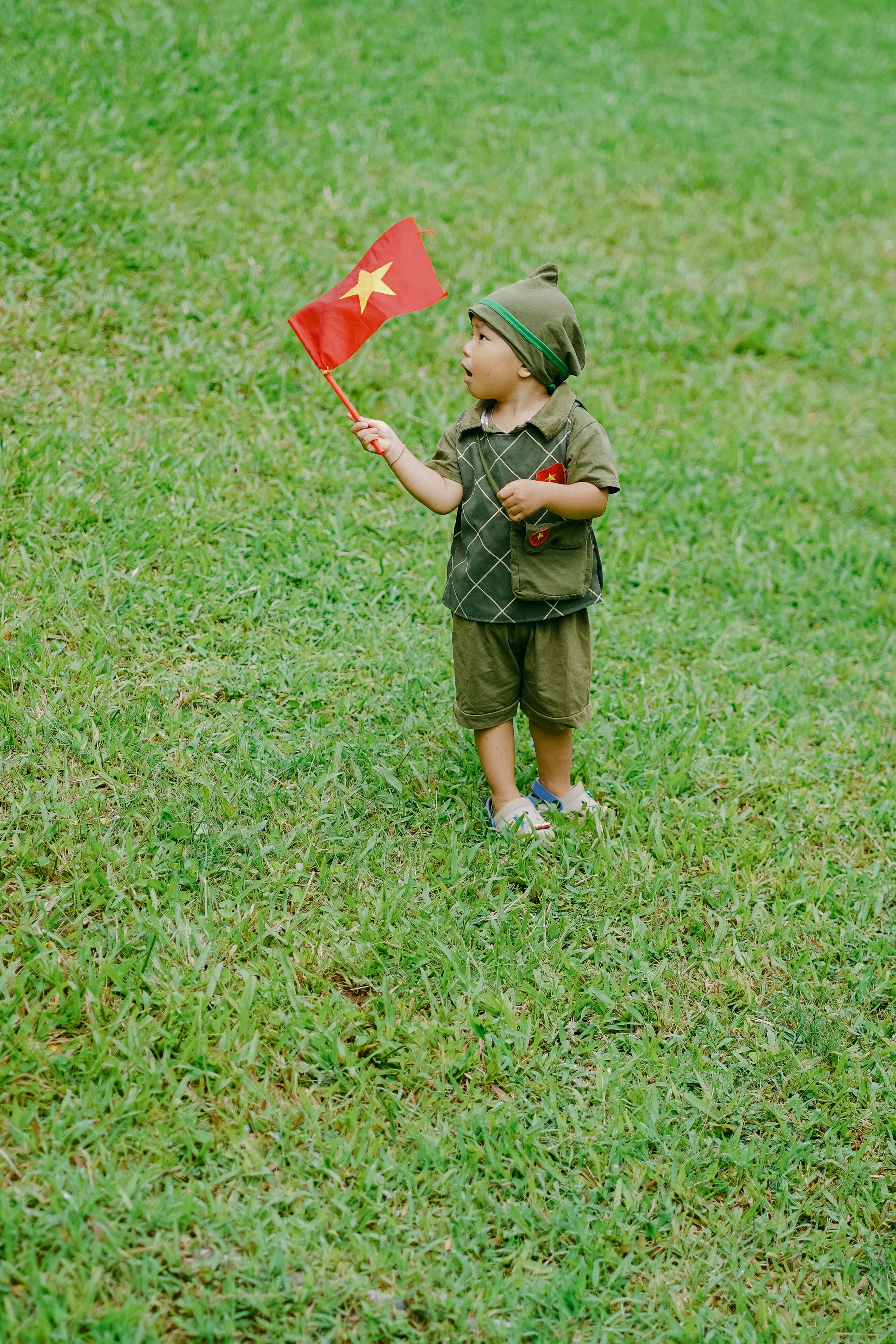 Young child in soldier costume waving a red star flag outdoors on a grassy field.