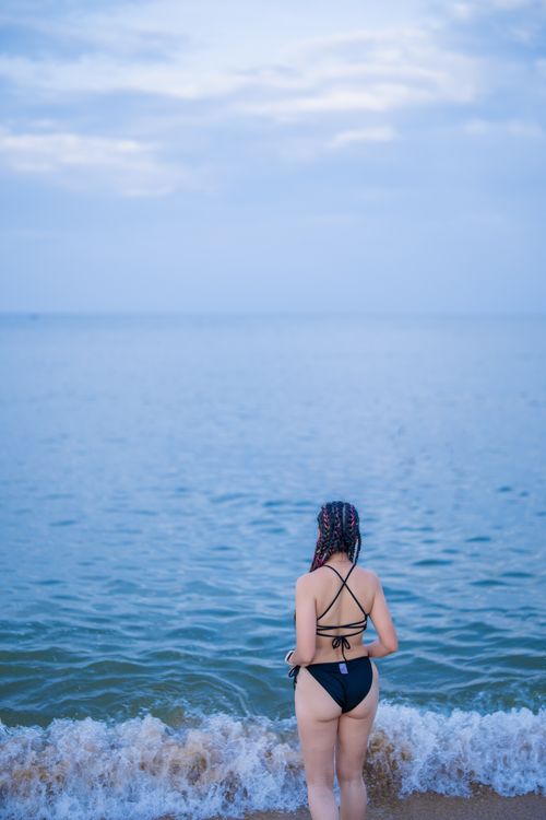 Free Peaceful scene of a woman in a black bikini standing at the beach, looking at the ocean. Stock Photo
