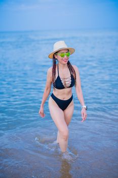 Confident woman in a black swimsuit and sunglasses walking along the beach in summer.