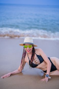 Woman in bikini and hat posing on beach with ocean backdrop.