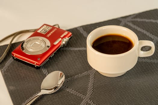 A white cup of coffee next to a red camera and spoon on a textured mat, perfect for stock use.