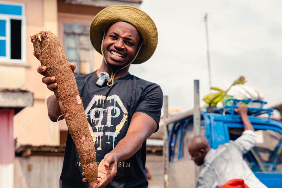 A smiling Ghanaian farmer holding up a large freshly harvested cassava root at a market, showing cassava as the dry-year anchor crop you can lift and sell when you need cash.