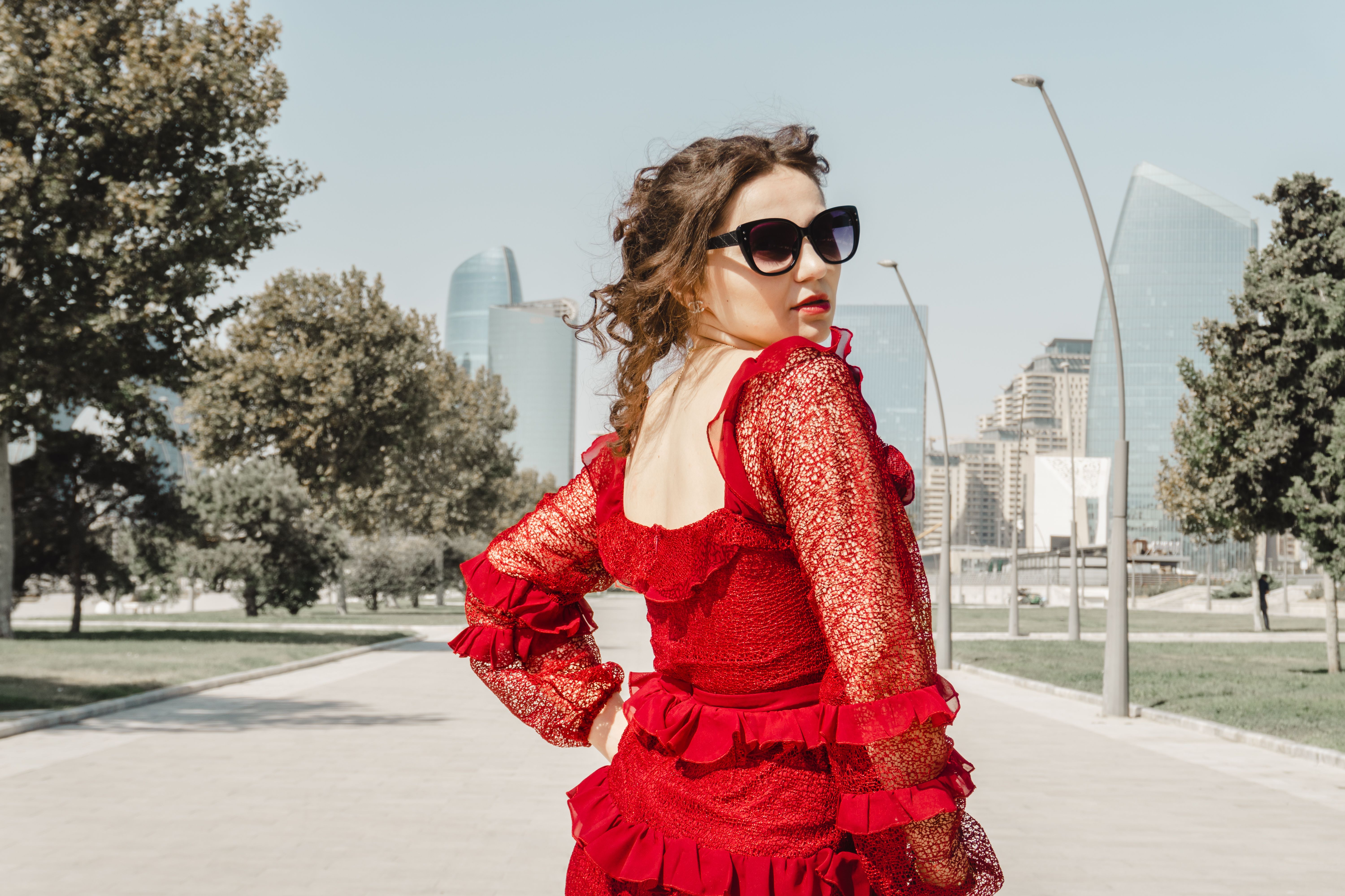 Une femme élégante vêtue d'une robe rouge avec des lunettes de soleil pose avec assurance à l'extérieur, avec des bâtiments modernes en arrière-plan.