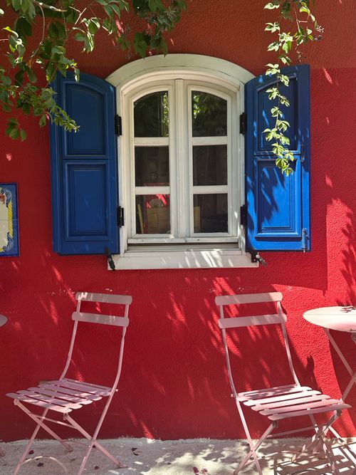 Free A bright red wall with a blue shuttered window and pink chairs exudes Mediterranean charm. Stock Photo