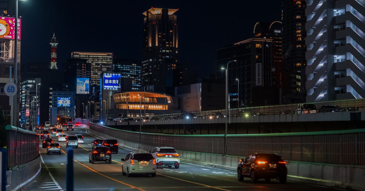 Cars driving on a highway at night with a vibrant city skyline in the background.