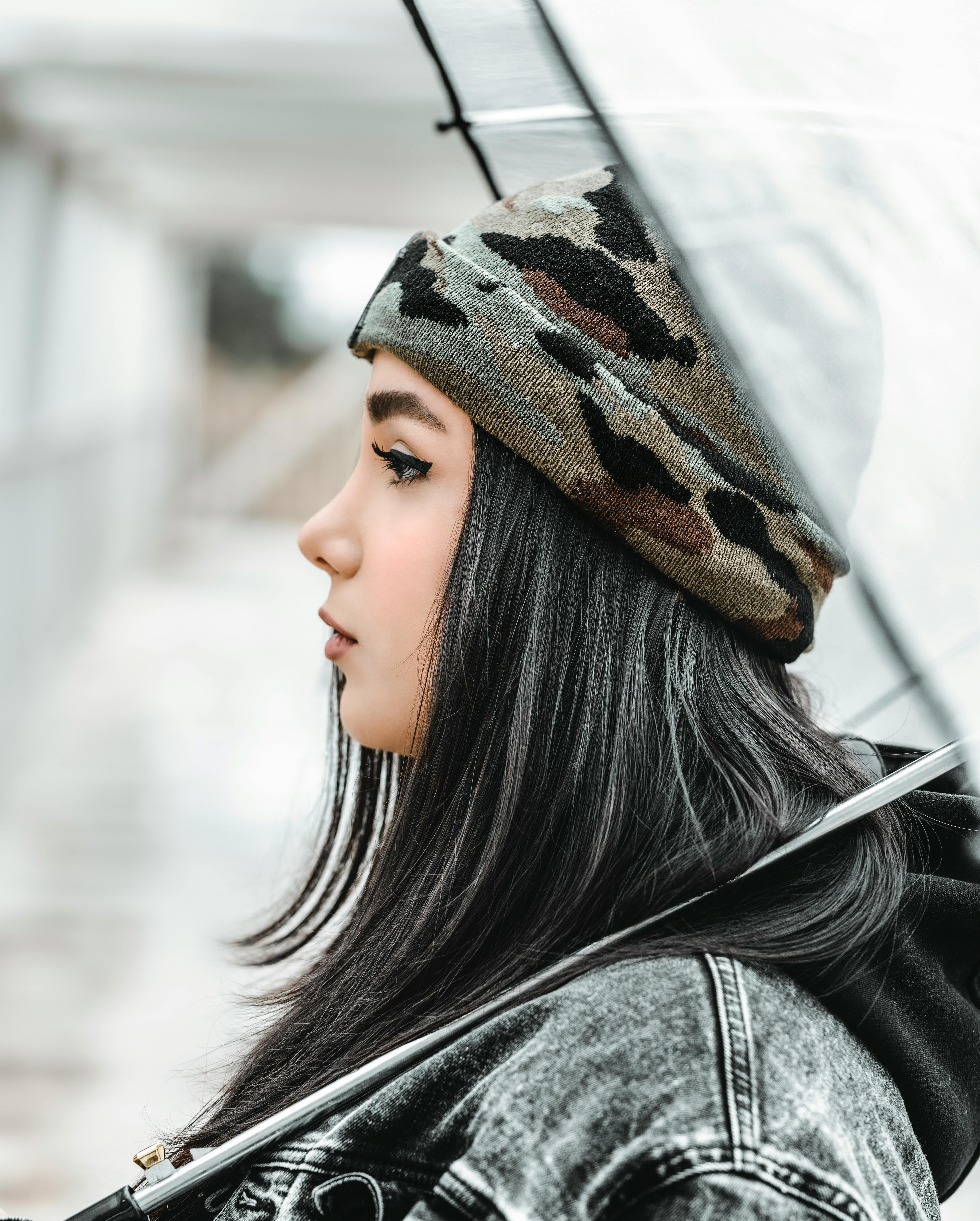 一个穿着牛仔夹克和迷彩帽的时髦妇女，在户外侧身拿着一把伞