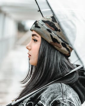 一个穿着牛仔夹克和迷彩帽的时髦妇女，在户外侧身拿着一把伞