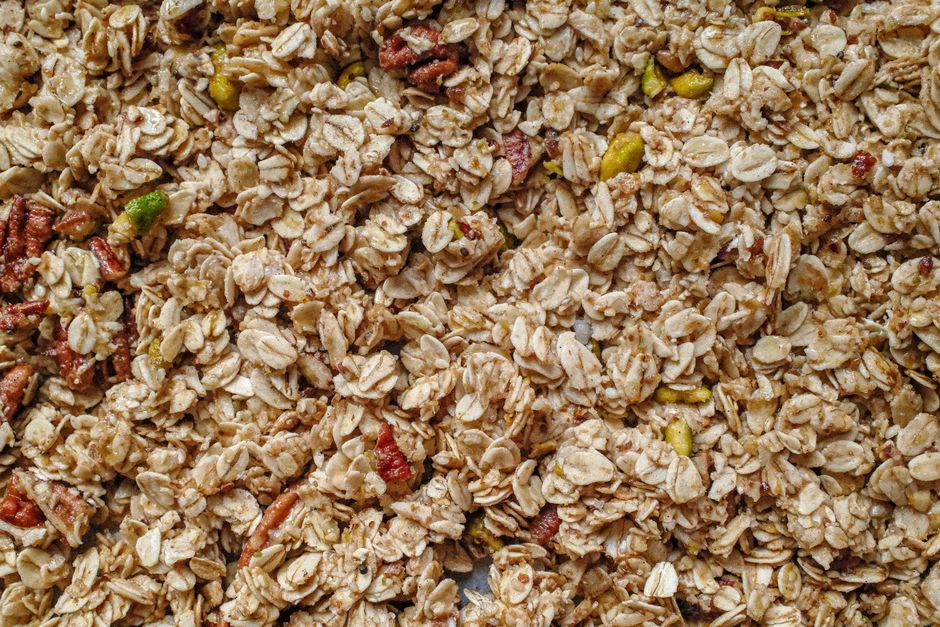 A sheet pan of golden baked gluten-free granola with clumpy clusters, almonds, and pumpkin seeds, next to a jar of the finished granola and a bowl with milk.