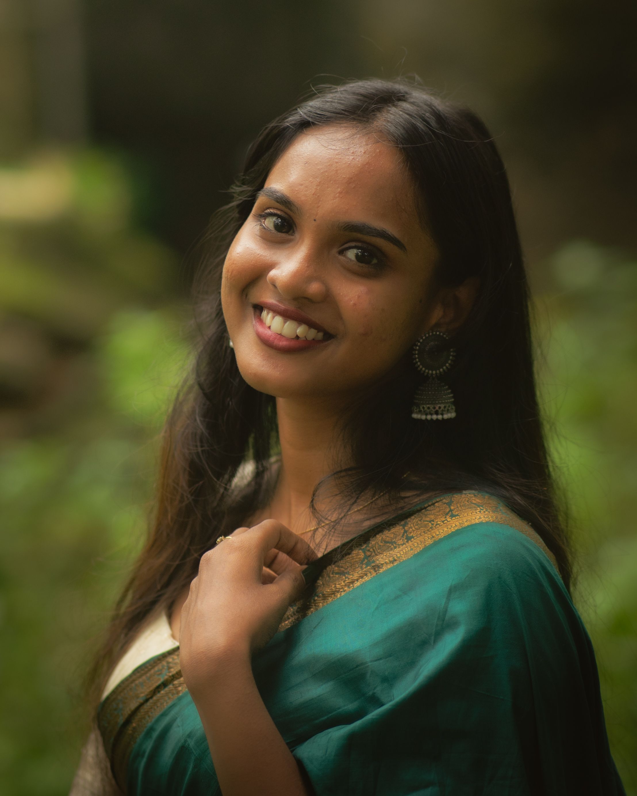 A young woman in an elegant green saree smiles warmly against a lush greenery background.