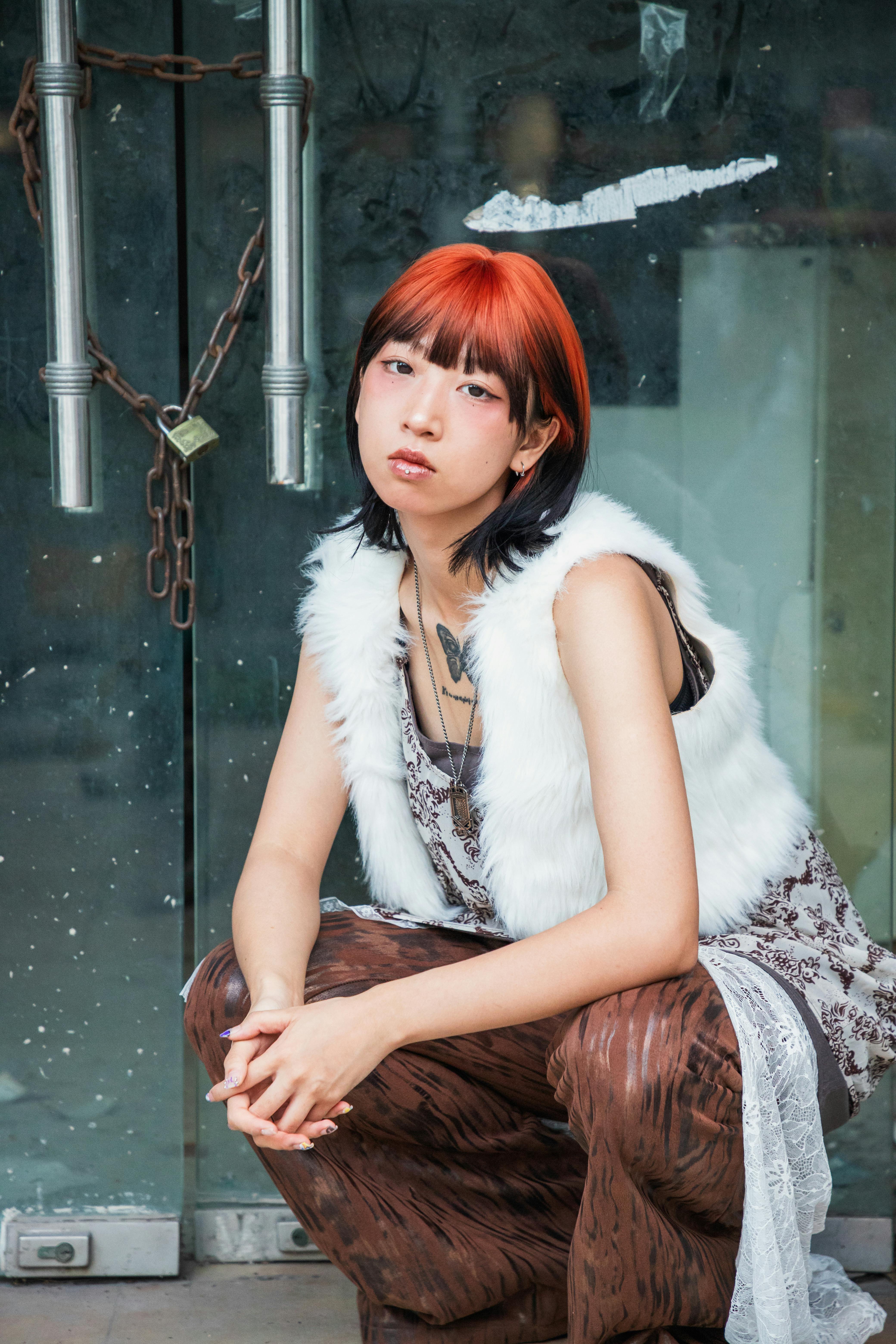 Ücretsiz Kızıl ve siyah saçlı, cesur moda anlayışına sahip şık genç kadın pencerenin önünde oturuyor. Kentsel moda havası. Stok Fotoğraflar