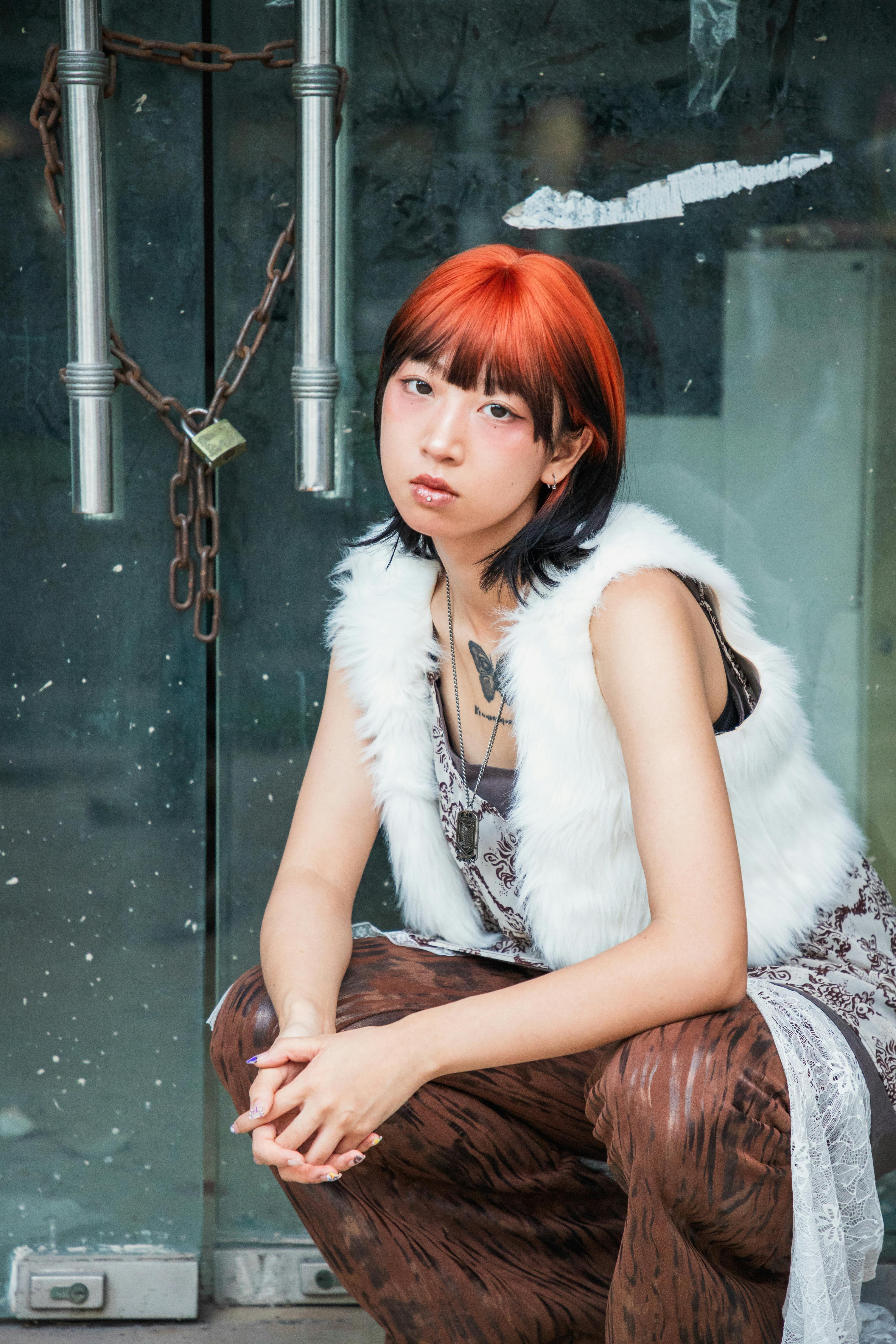 Free Trendy woman with striking red hair poses stylishly with urban backdrop. Stock Photo