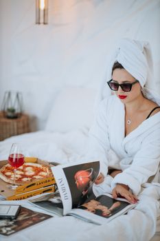 Woman in robe enjoying pizza and magazines in bed for a relaxed morning moment.