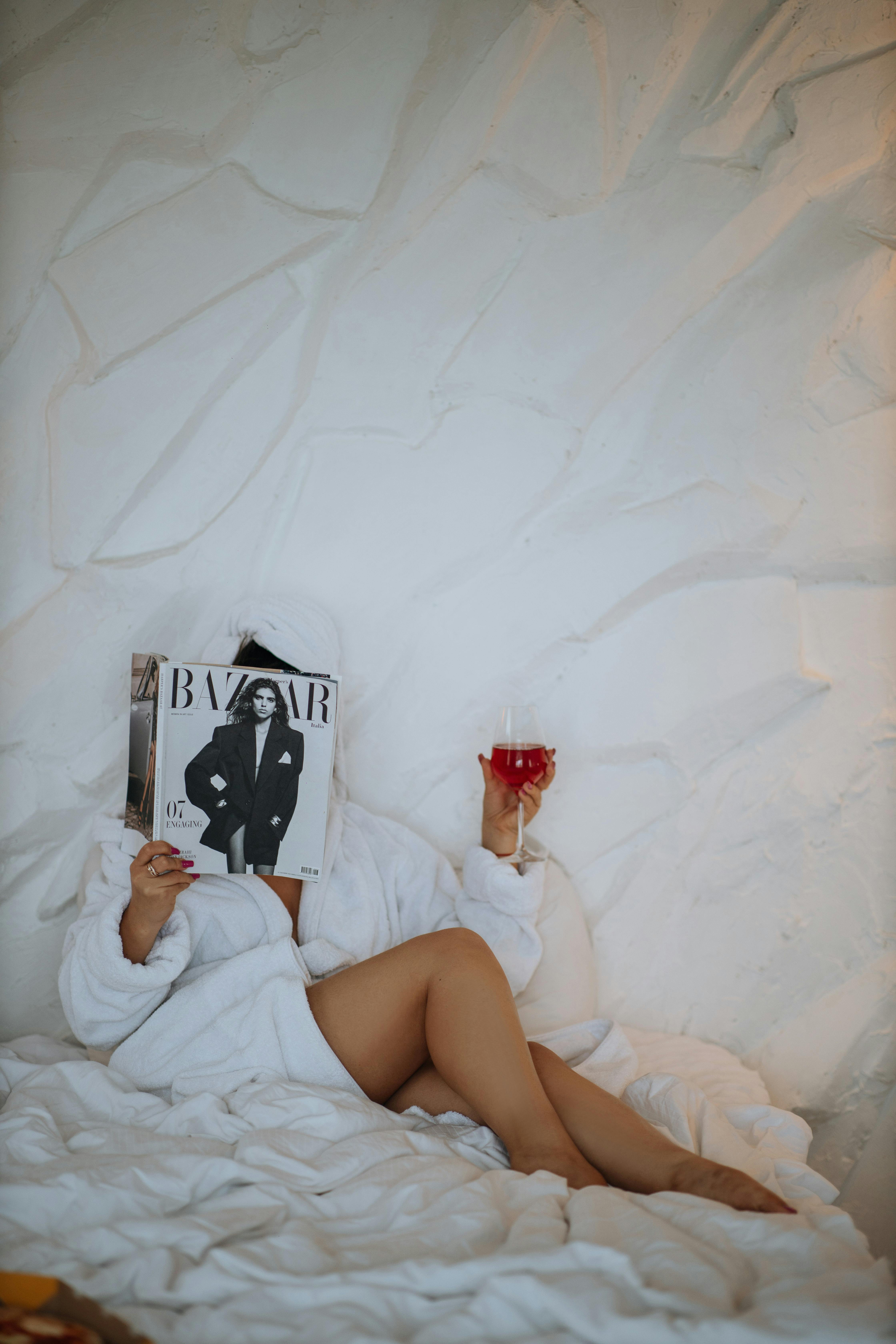 A woman in a white robe relaxes on a bed while reading a fashion magazine and holding a drink.