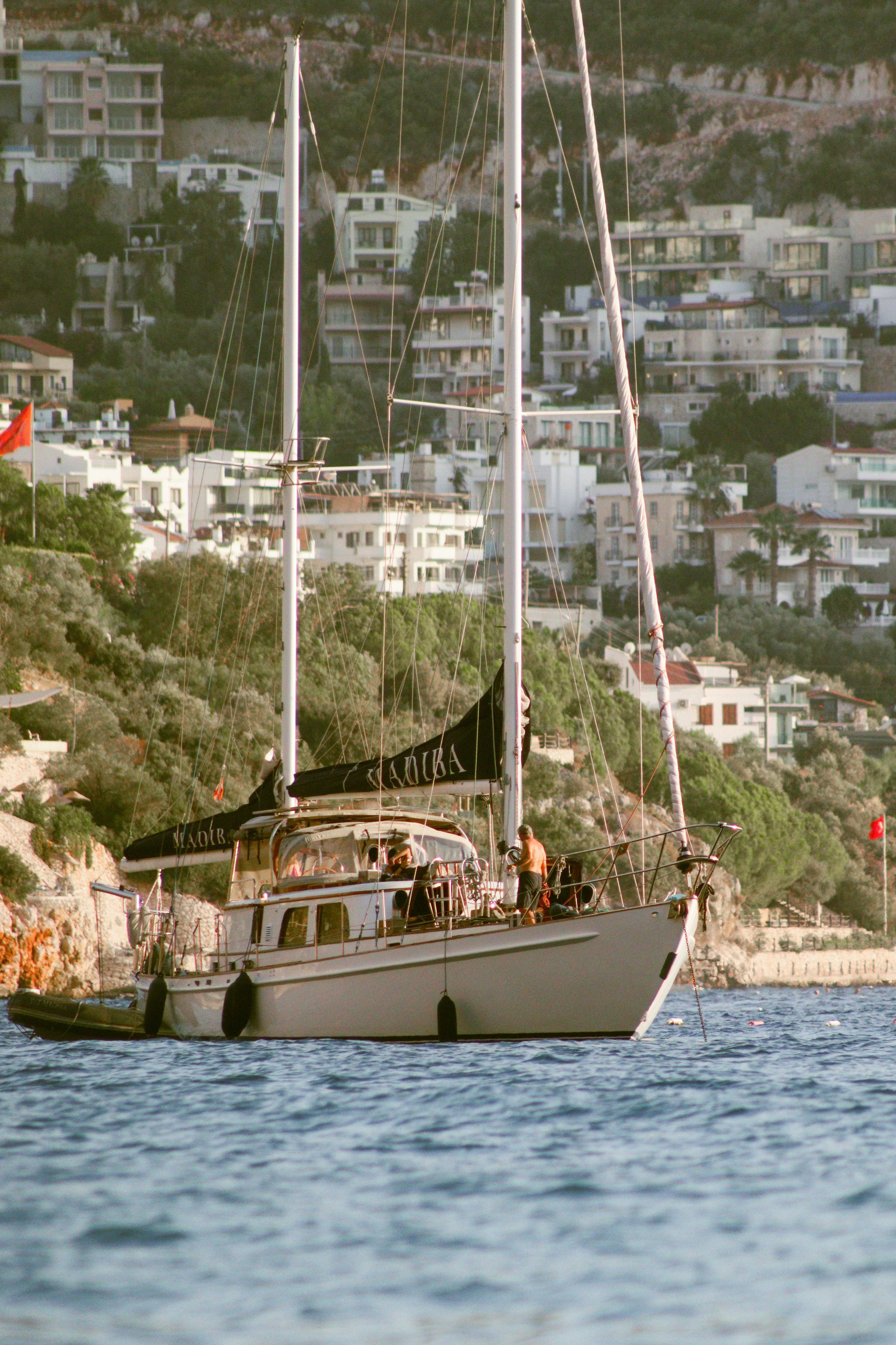 A sailing yacht anchored close to a coastal city with lush greenery and modern buildings under a sunny day.