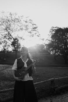 Black and white photo of a woman with a ukulele, outdoors at sunset.