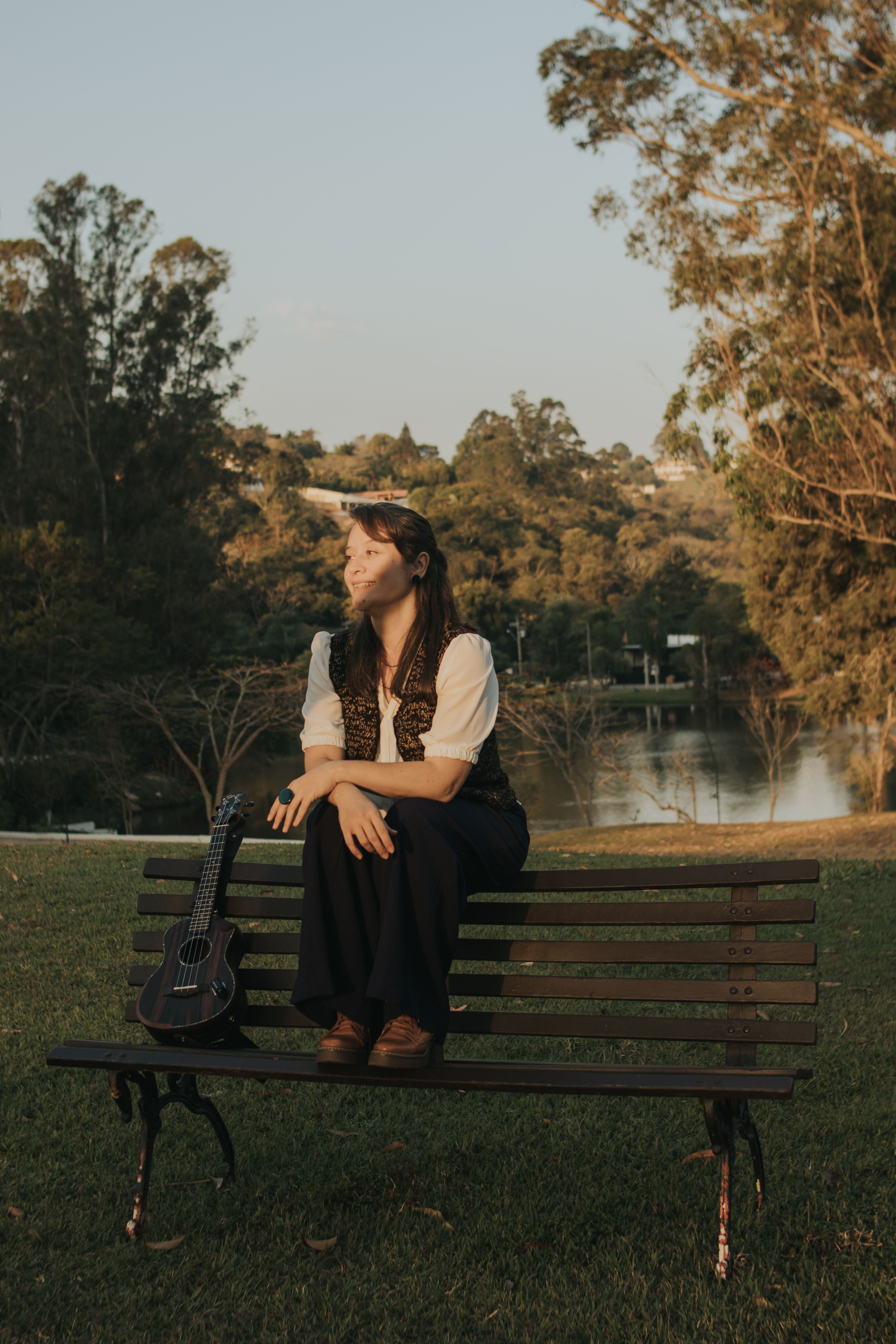 A young woman sits on a bench in a park with a ukulele, enjoying the outdoor scenery.