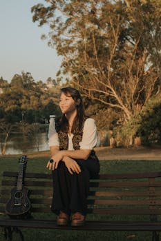 Smiling woman sitting on a bench with a ukulele during sunset in a tranquil park.