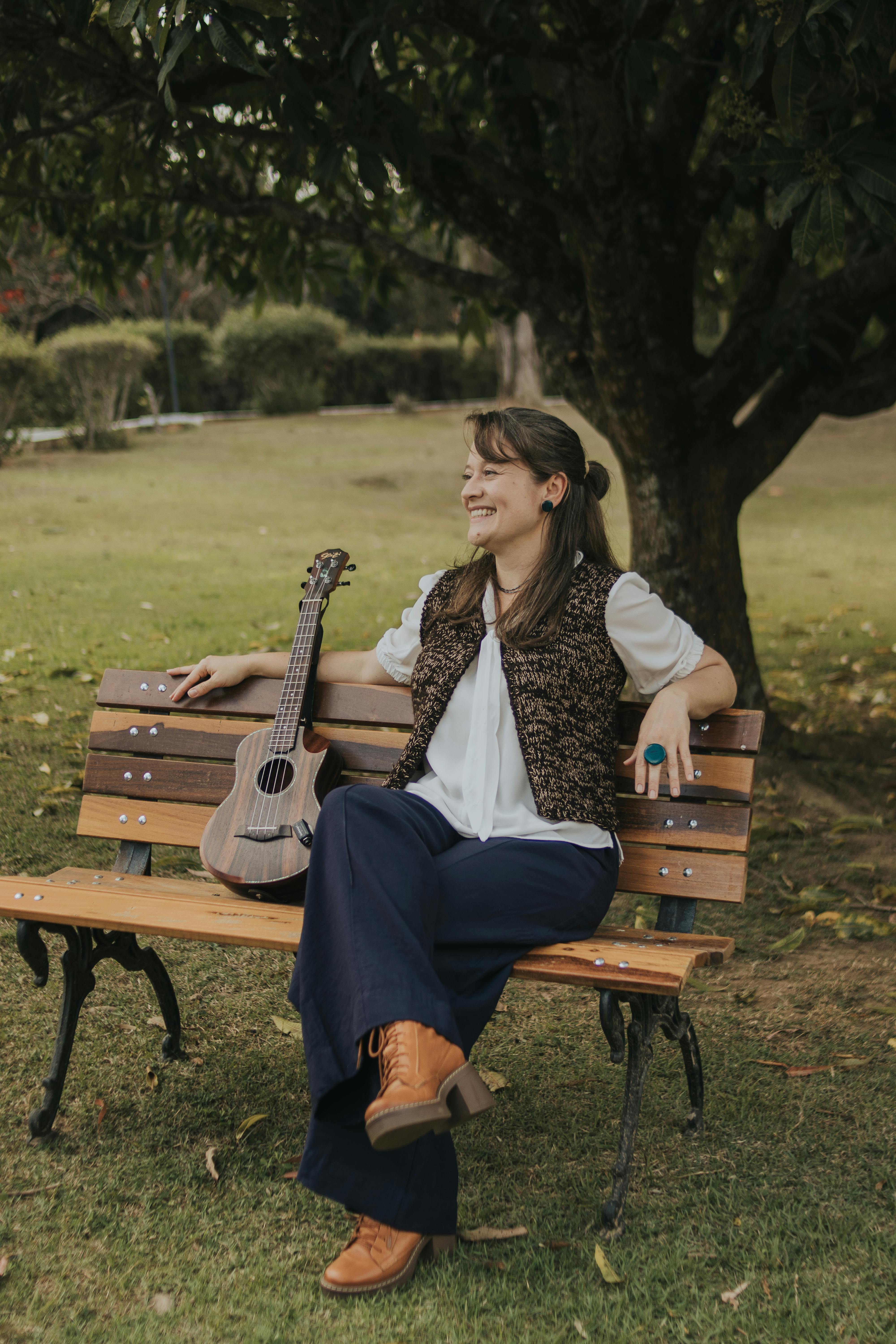 Cheerful woman sitting on park bench with ukulele, enjoying a sunny day outdoors.