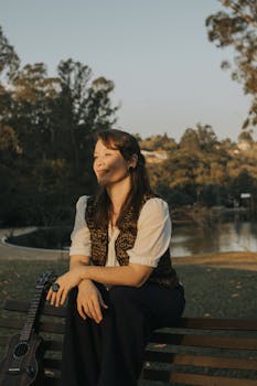 A woman enjoys a peaceful moment on a park bench with a guitar at sunset.