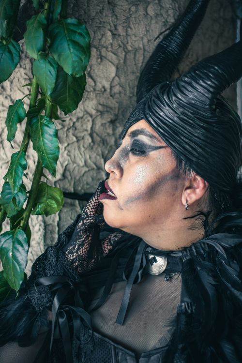 Free A person in creative gothic attire and makeup, featuring horns, posing outdoors next to leafy plants. Stock Photo