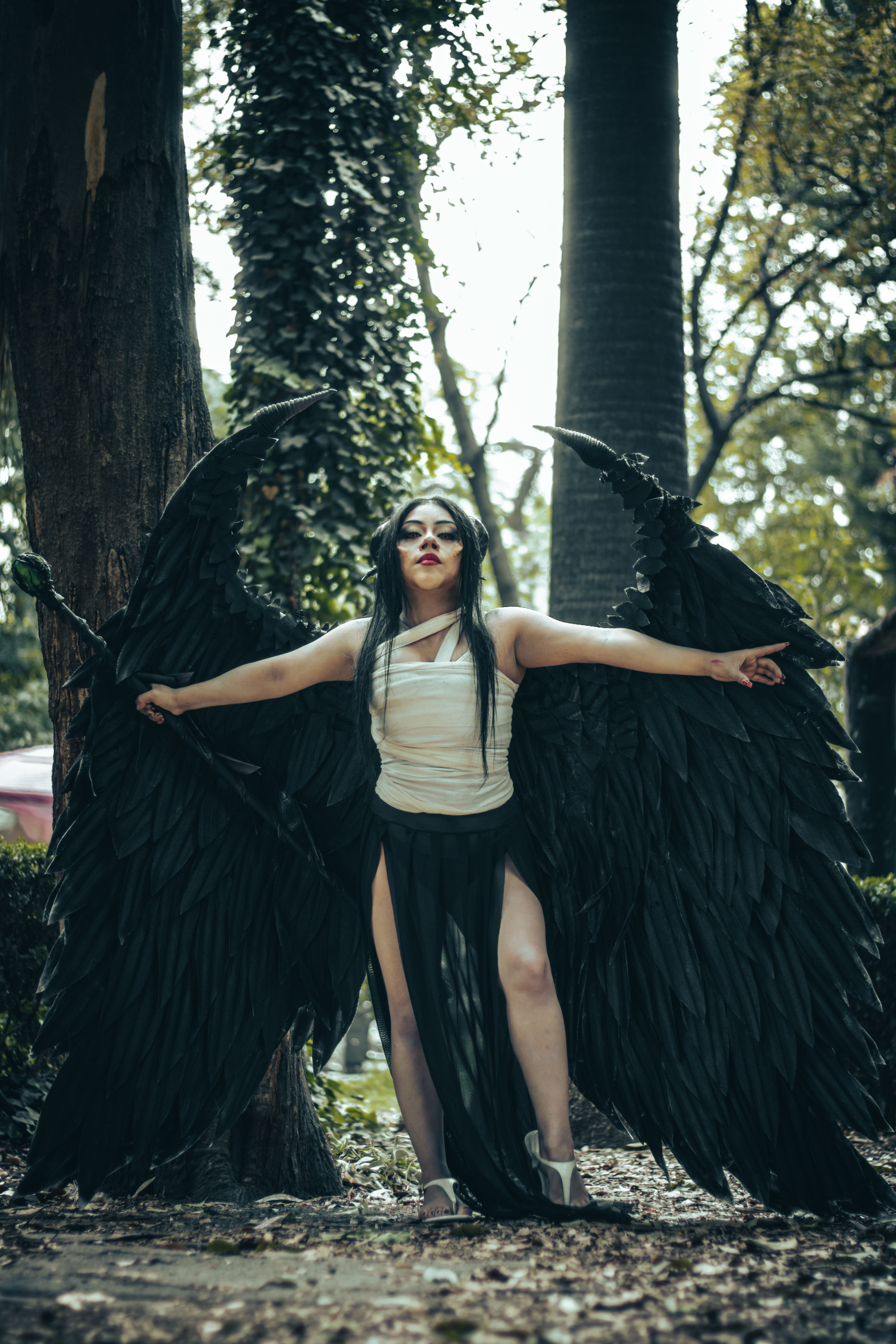 A person with black wings in artistic cosplay posing outdoors in a park.