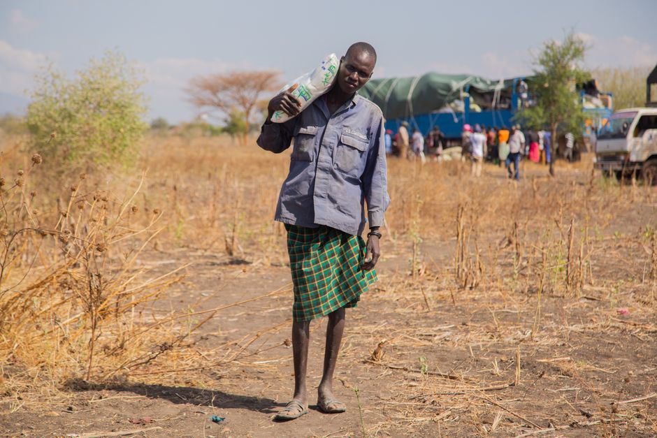 A man carries a bag in a rural African landscape, with a truck and people in the background.