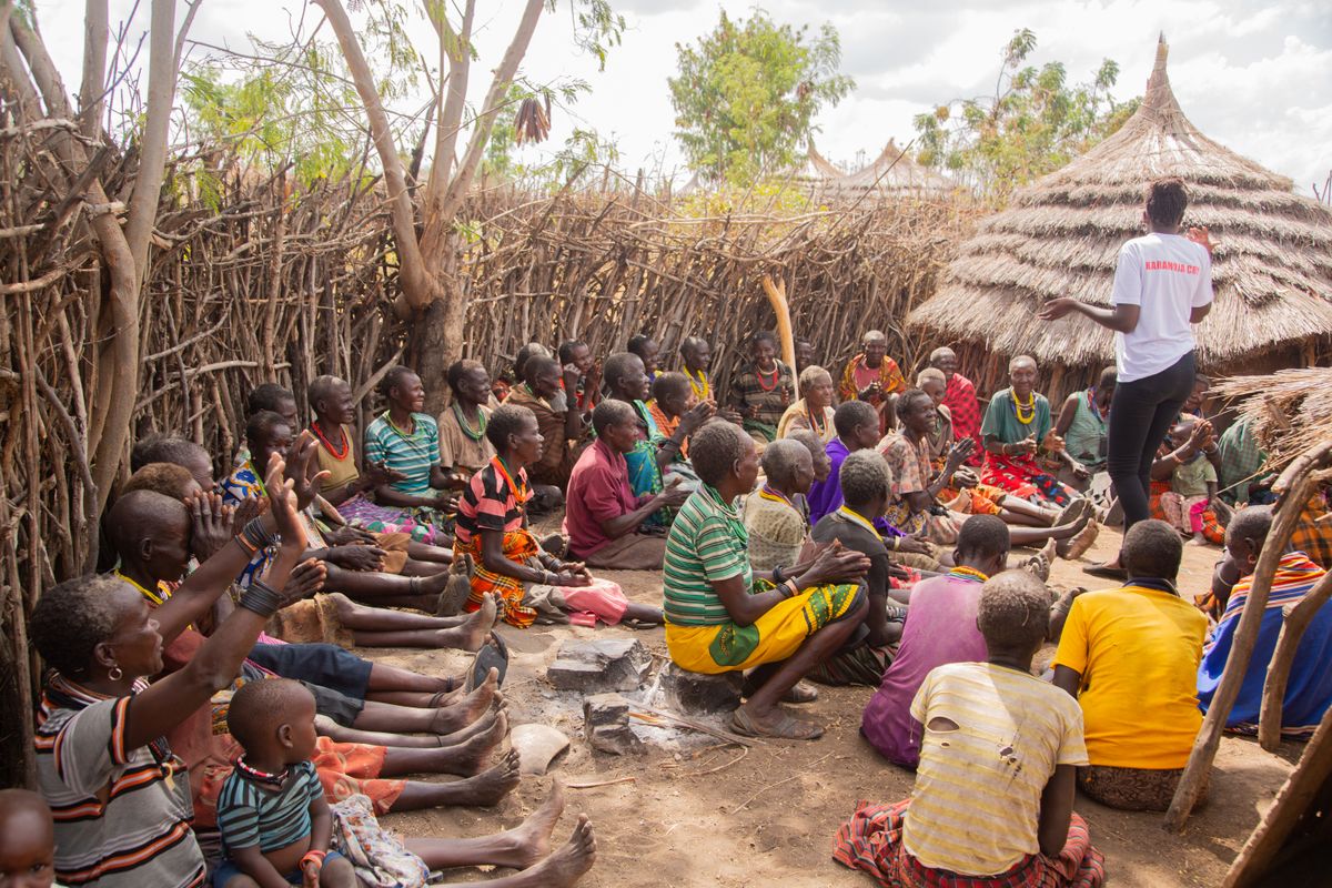 Community members gather outdoors in a rural village, seated together in conversation.