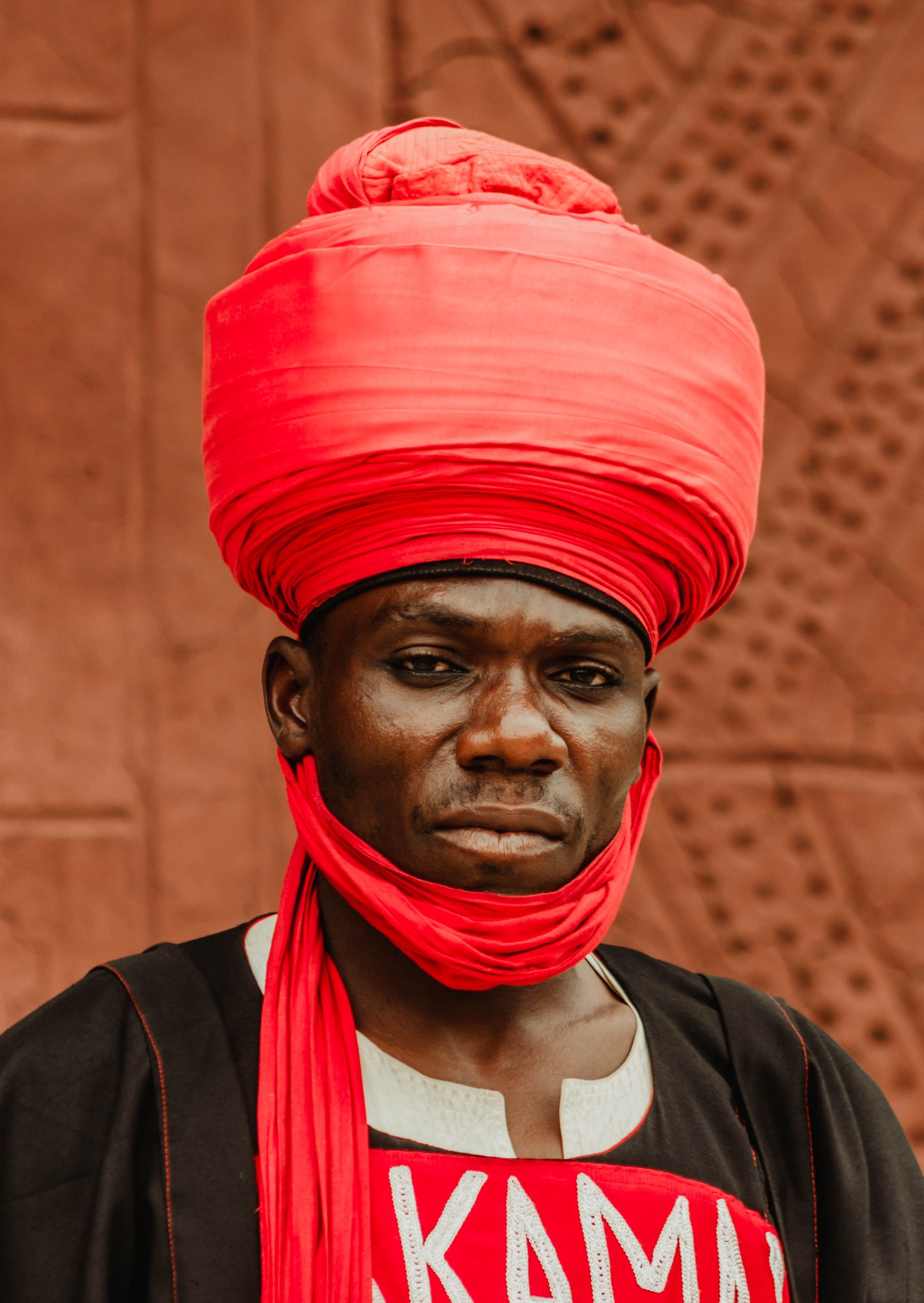 A striking portrait of a man in a traditional red headwrap and attire against a textured backdrop.