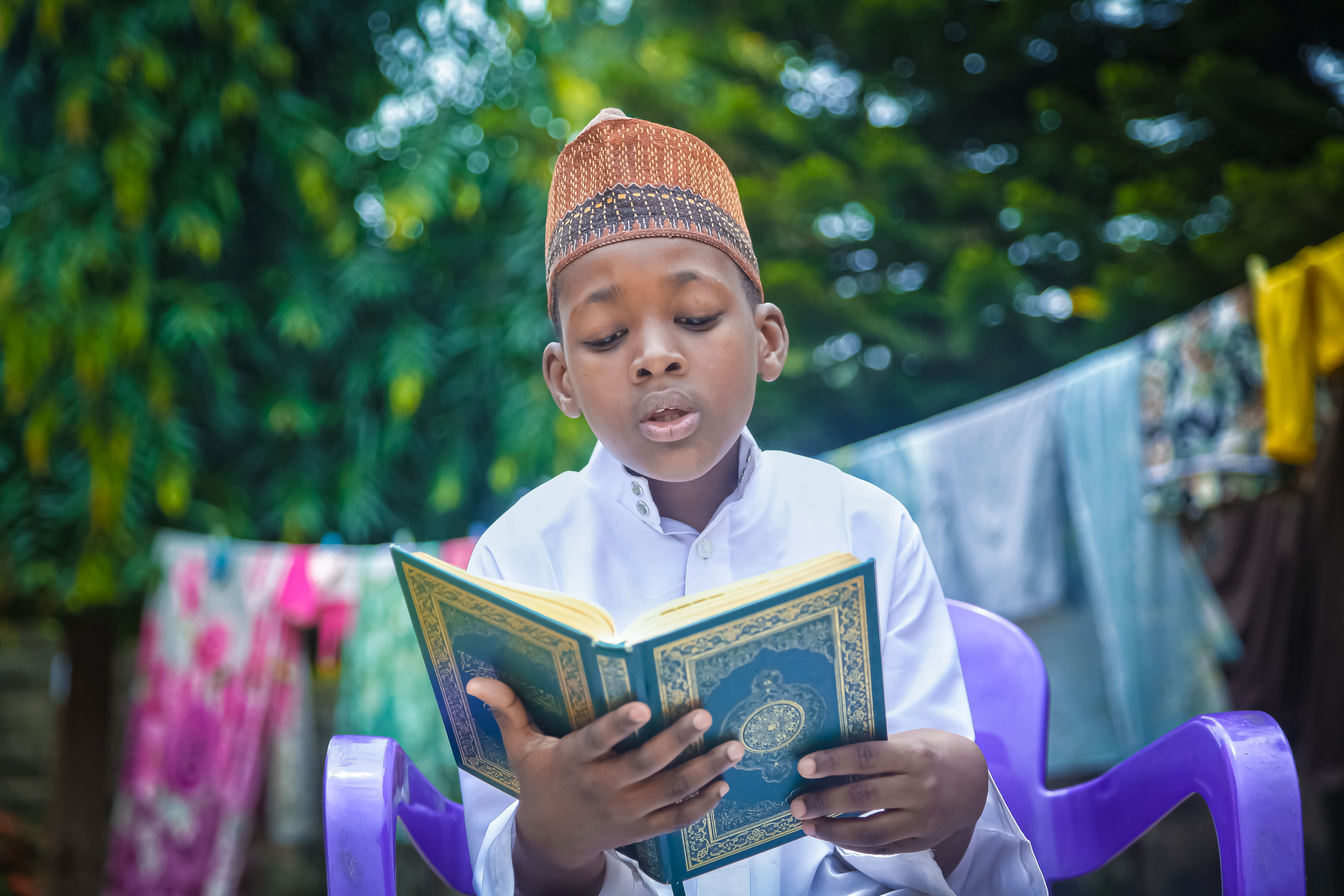 A young boy in traditional attire reads the Quran outdoors, surrounded by nature.