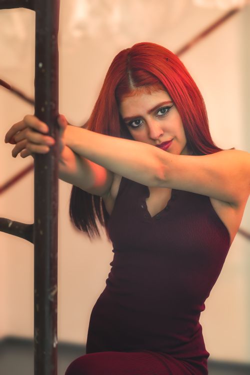 Free Elegant portrait of a woman with striking red hair, leaning on a pole indoors. Stock Photo