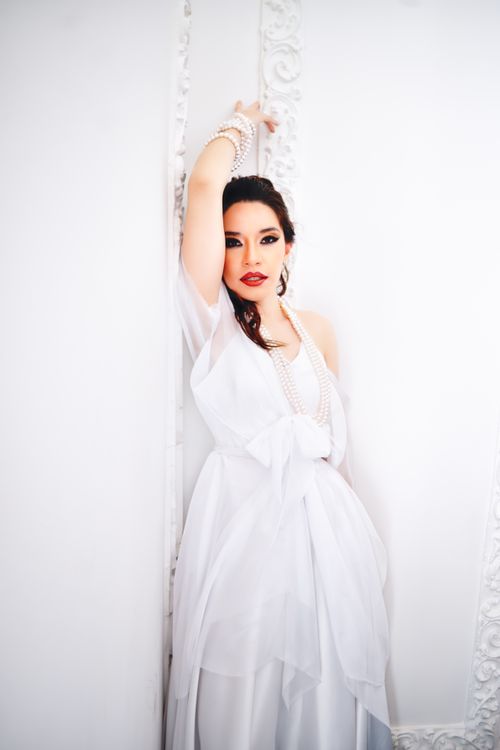 Free Stylish woman in flowing white dress with pearl jewelry posing indoors. Stock Photo