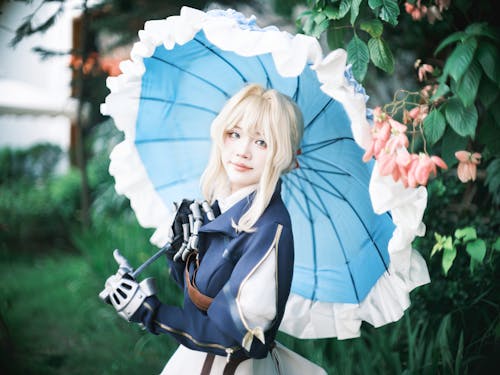 Woman in a detailed costume holding a blue parasol, surrounded by vibrant green foliage.