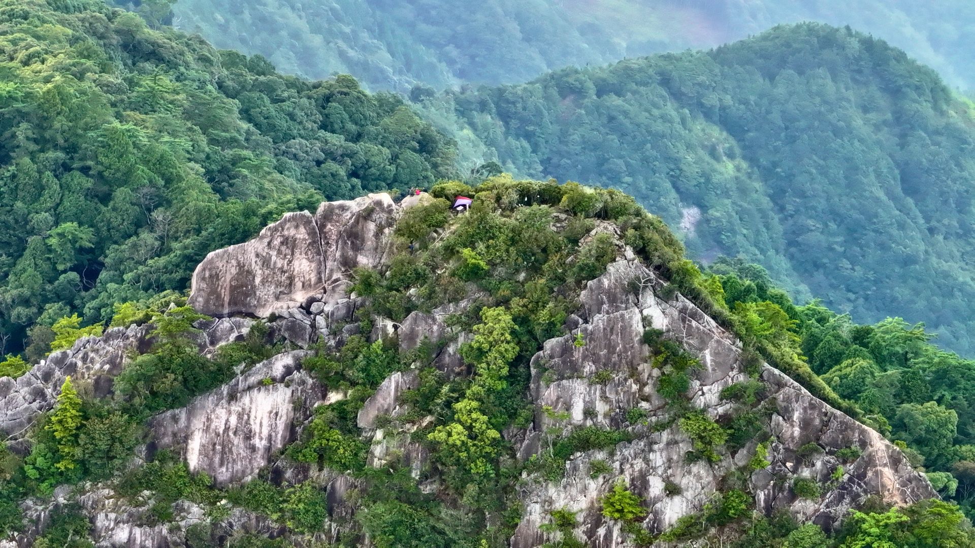 A stunning aerial view of a lush, green mountain peak surrounded by dense forest and rocky formations.