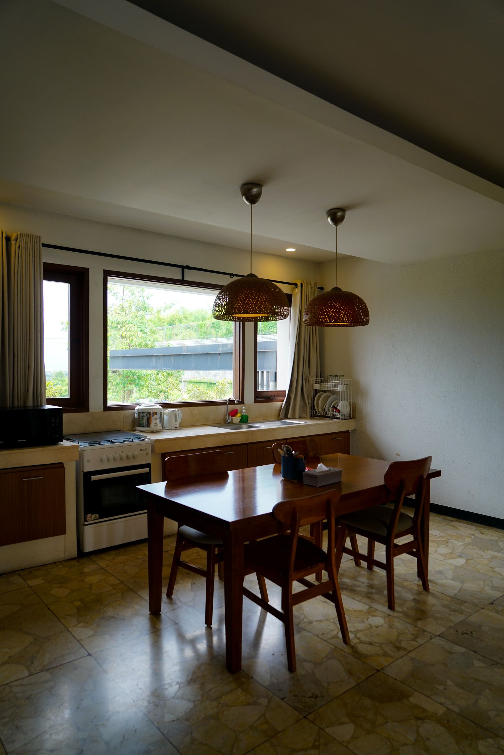 Wood dining table and curved-back chairs beneath two woven pendants beside a kitchen window