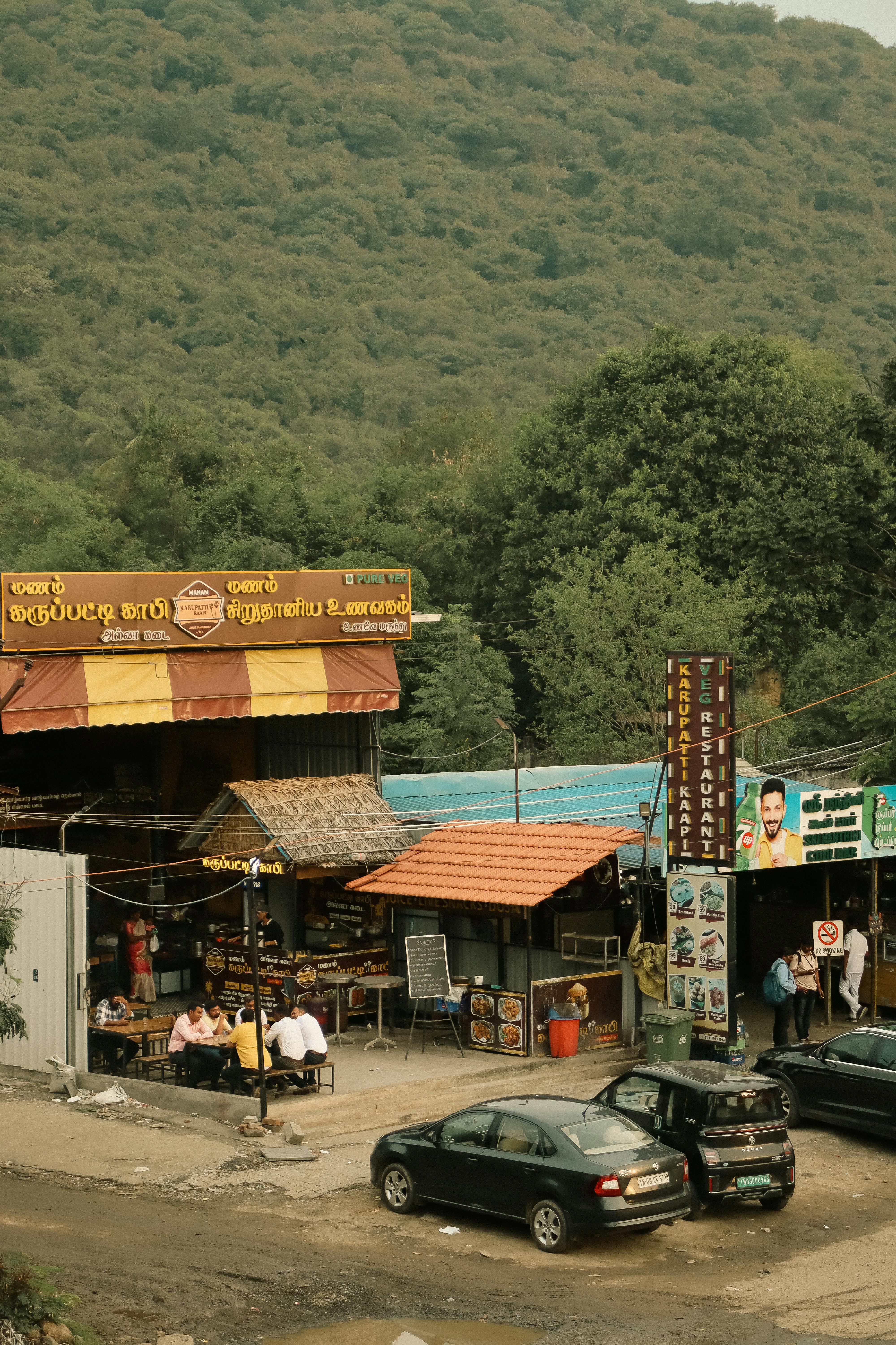 Street view of a local market with cars and people against a scenic mountain backdrop.