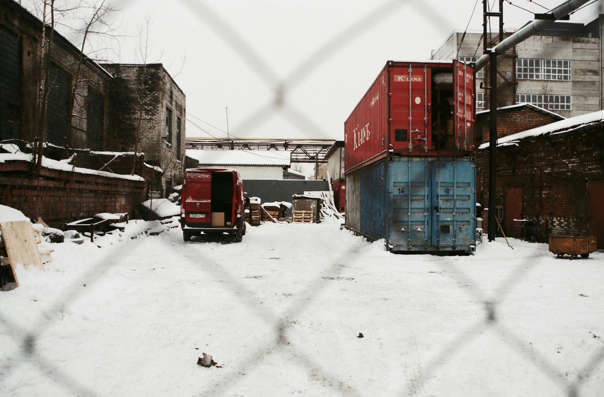 Snow-covered industrial yard with shipping containers, behind chain link fence.