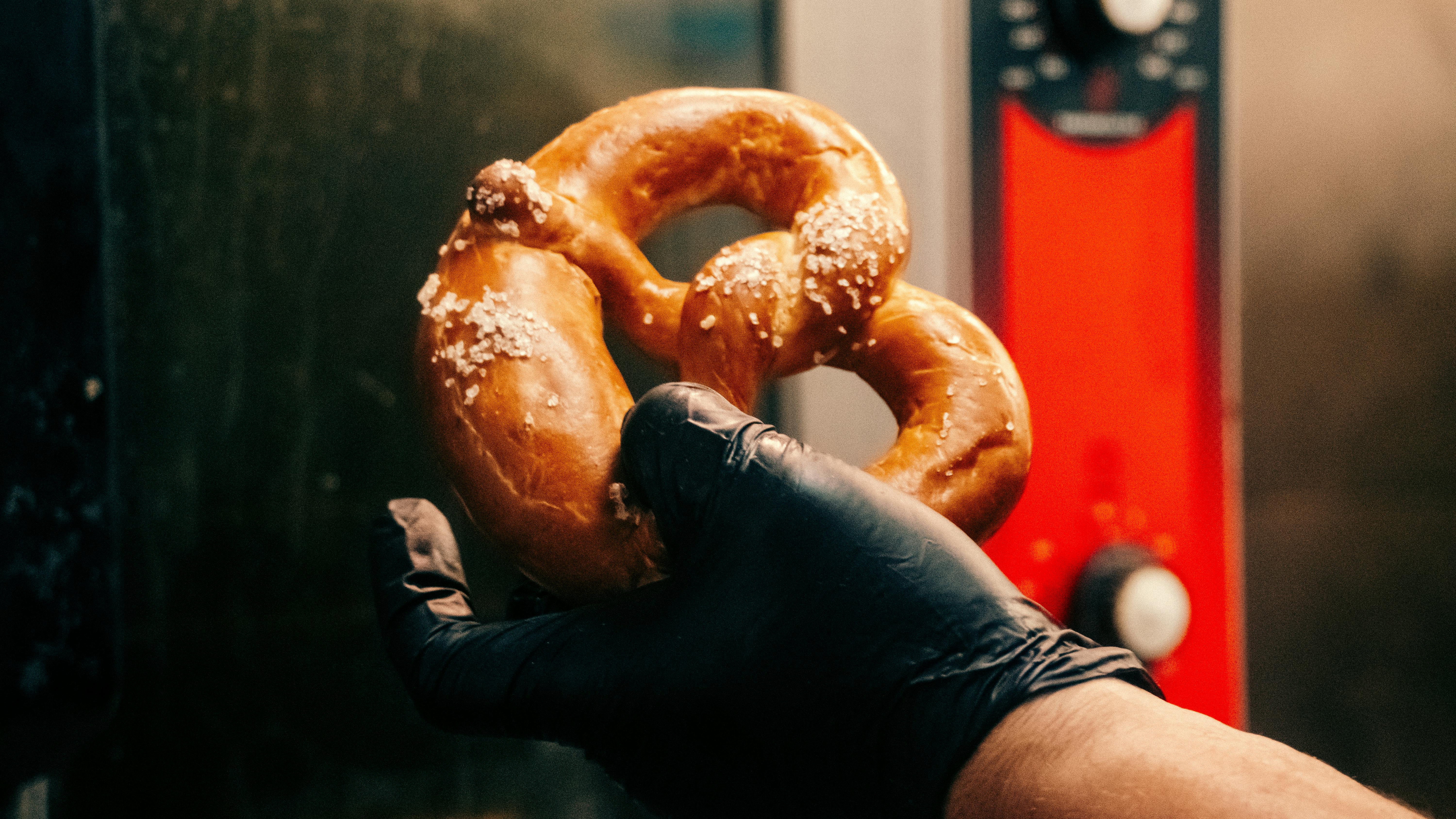 A black-gloved hand holding a freshly baked pretzel with coarse salt in a kitchen setting.