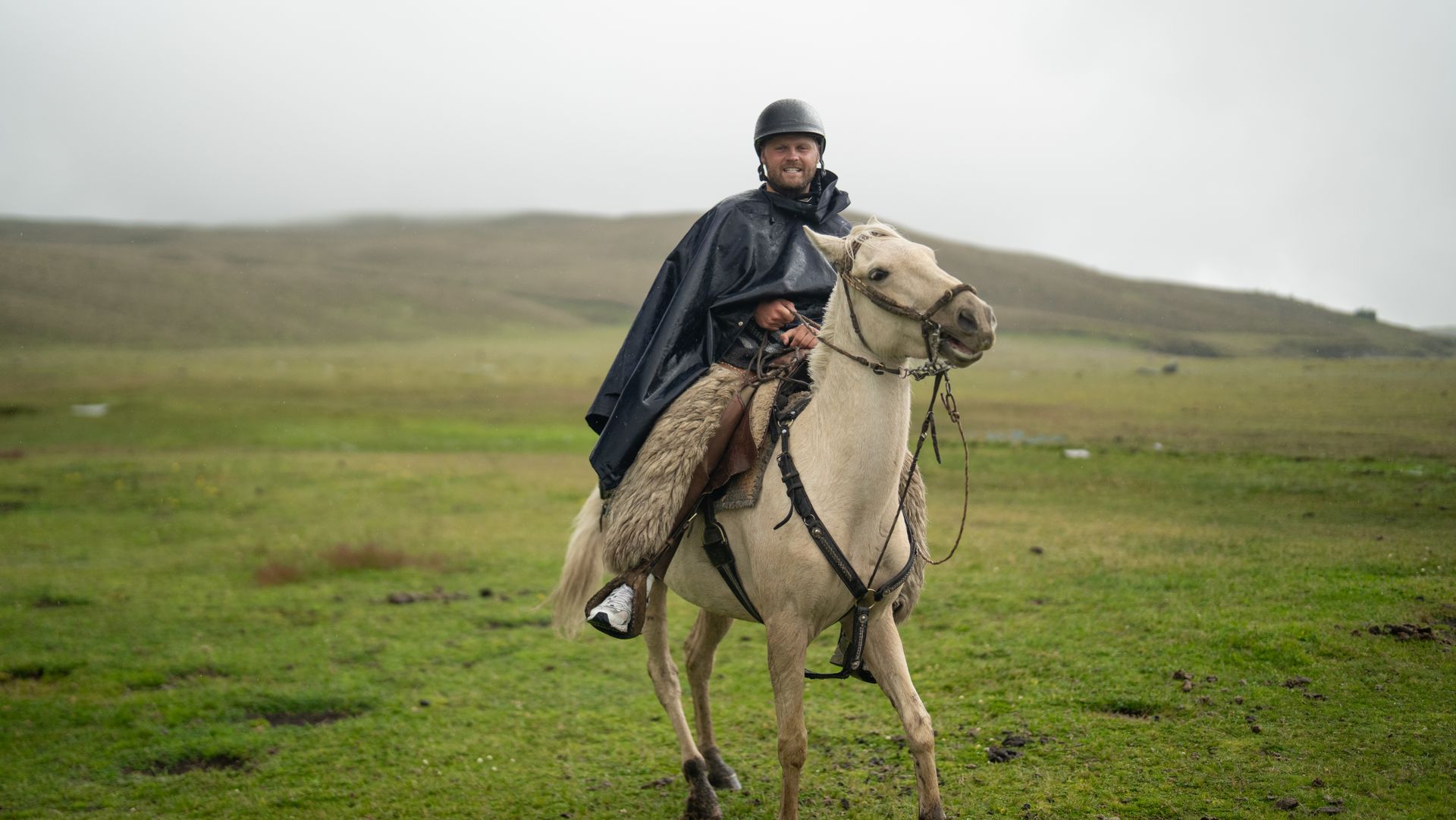 A man in a raincoat rides a horse across a grassy field on a cloudy day.
