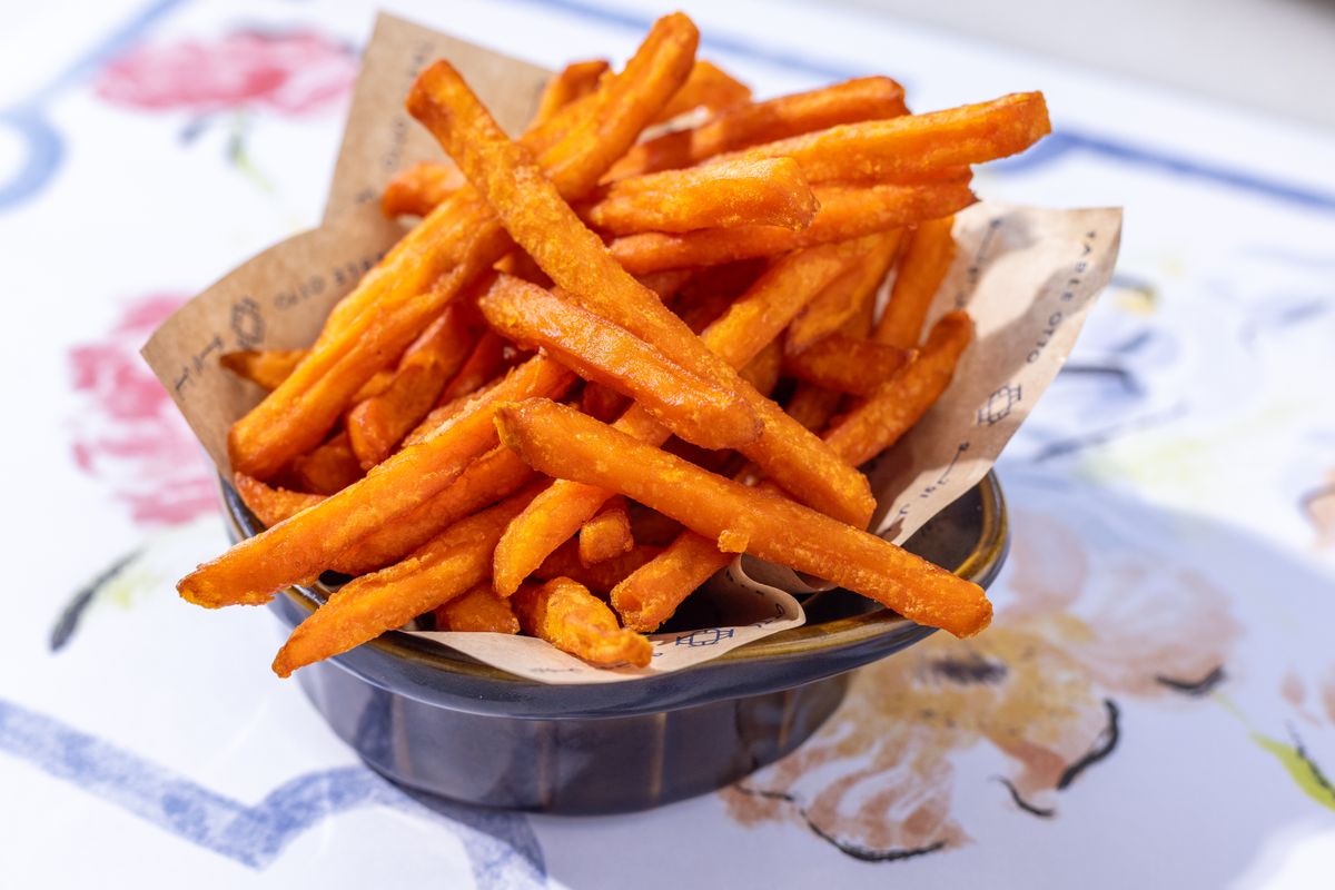 Thin orange sweet potato fries piled in a paper-lined dark ceramic bowl on a floral tablecloth