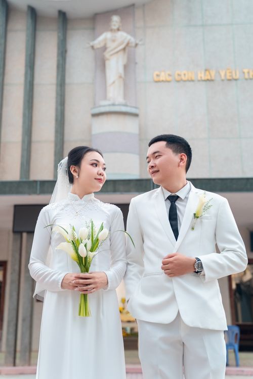 Free A bride and groom in white outfits stand together in front of a church, gazing at each other lovingly. Stock Photo