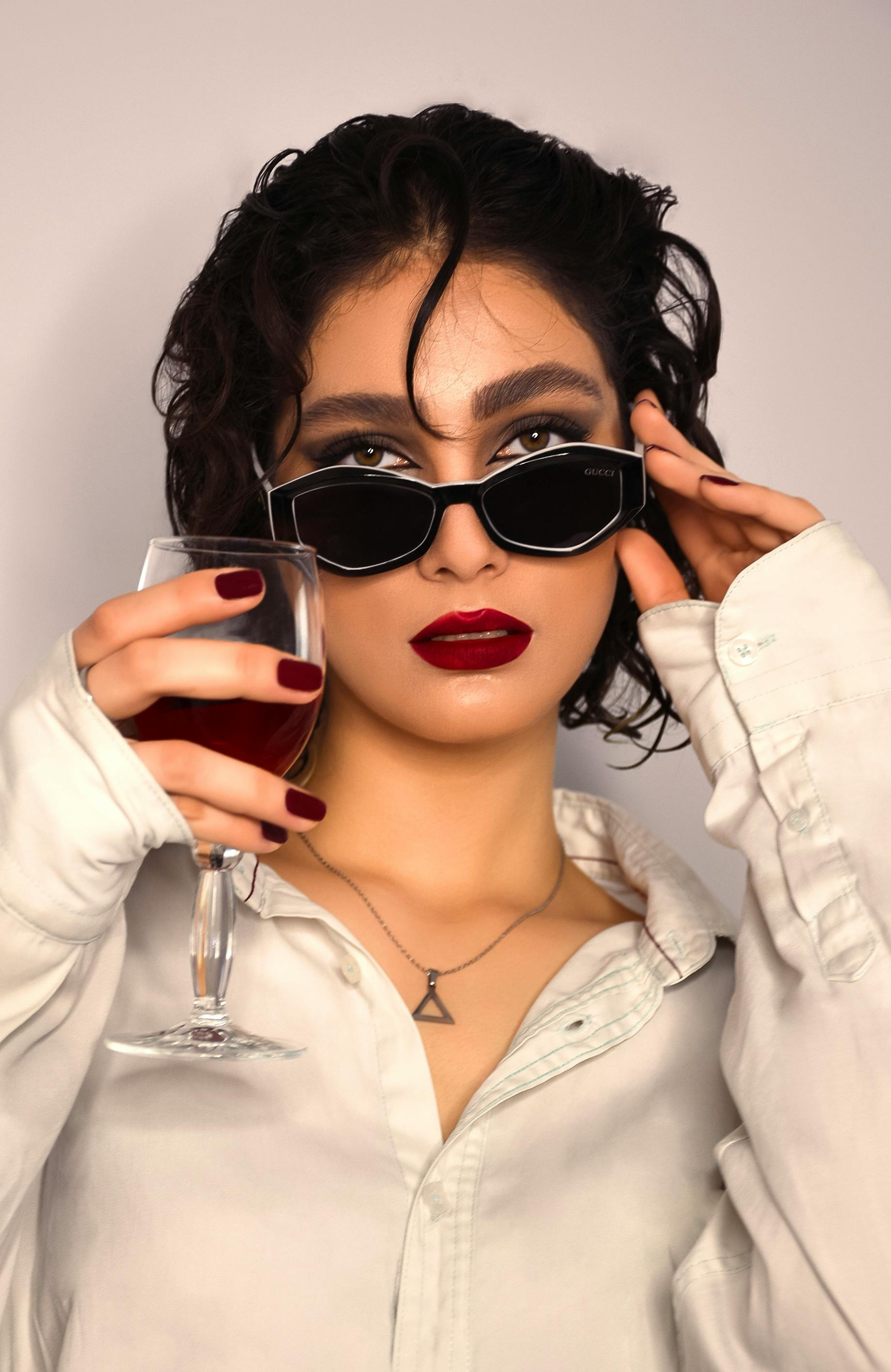 Elegant woman in sunglasses holding red wine, exuding modern style.