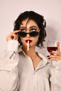 Fashionable woman in sunglasses holding wine and cigarette indoors.
