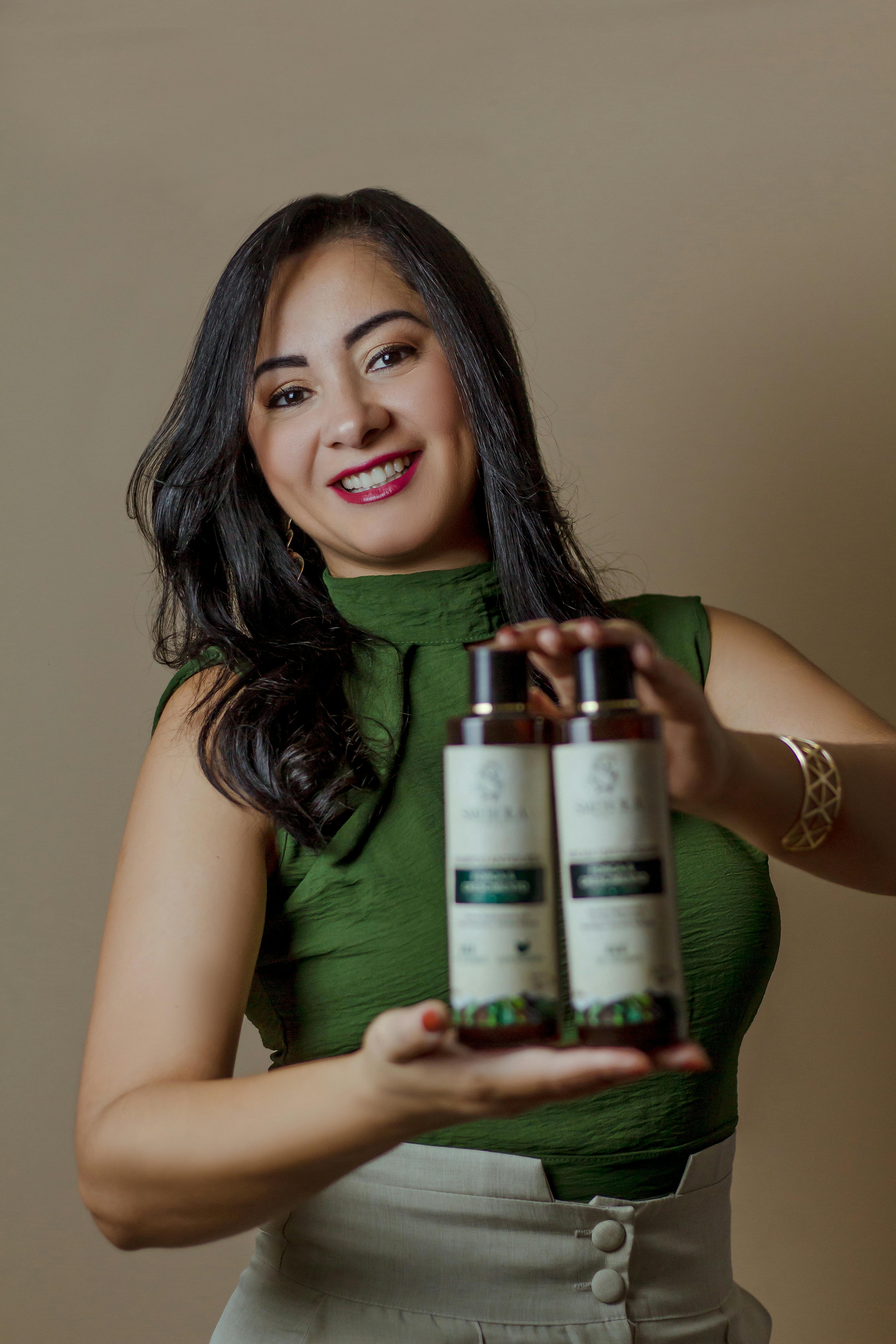 Smiling woman in green dress holds two bottles of organic hair products against beige background.