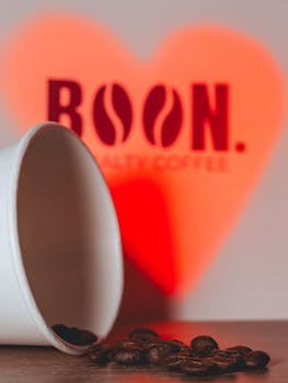 A paper cup spilling coffee beans on a wooden surface with a glowing heart-shaped BOON logo background.