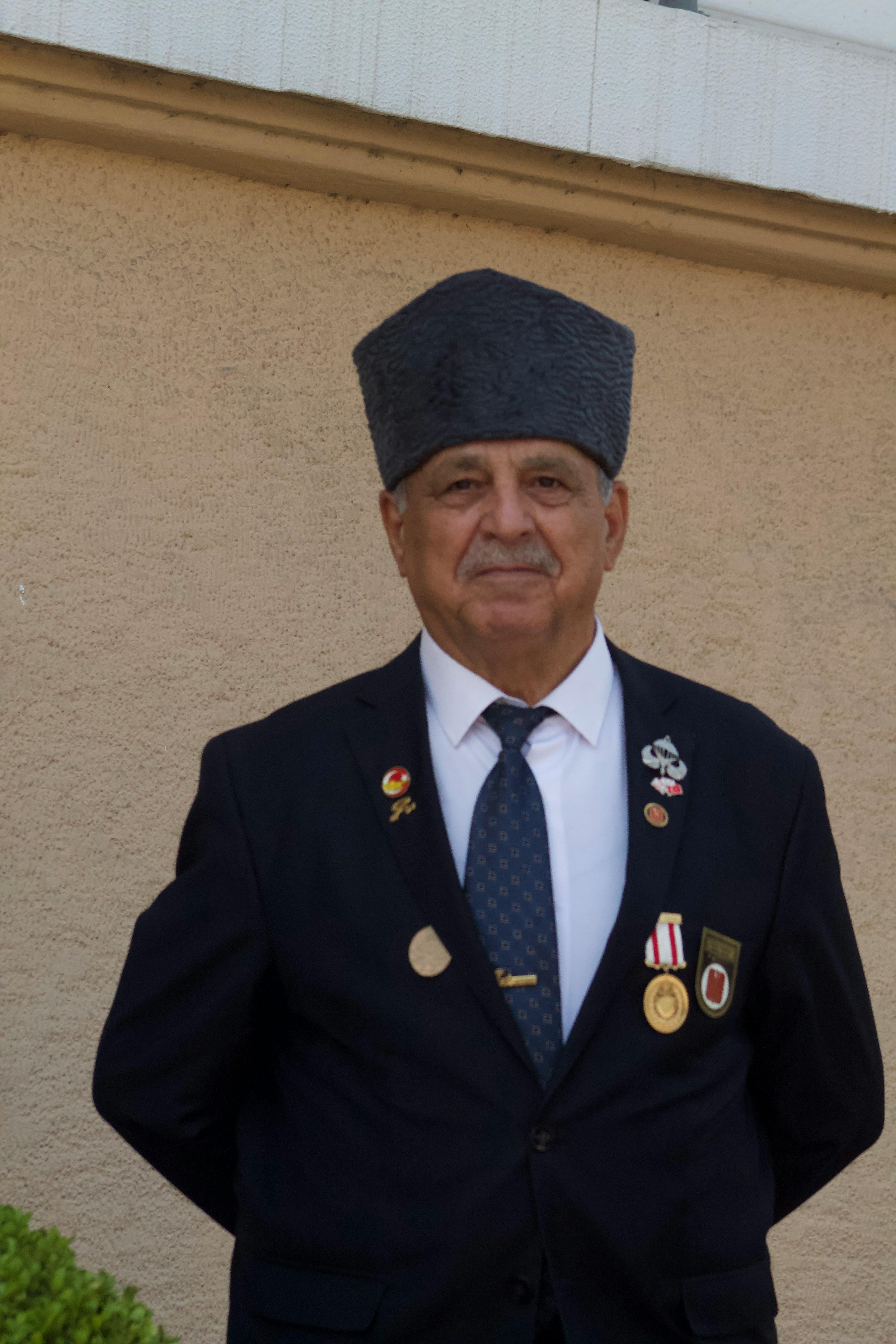Elderly gentleman wearing a traditional hat, medals on suit, standing outdoor.