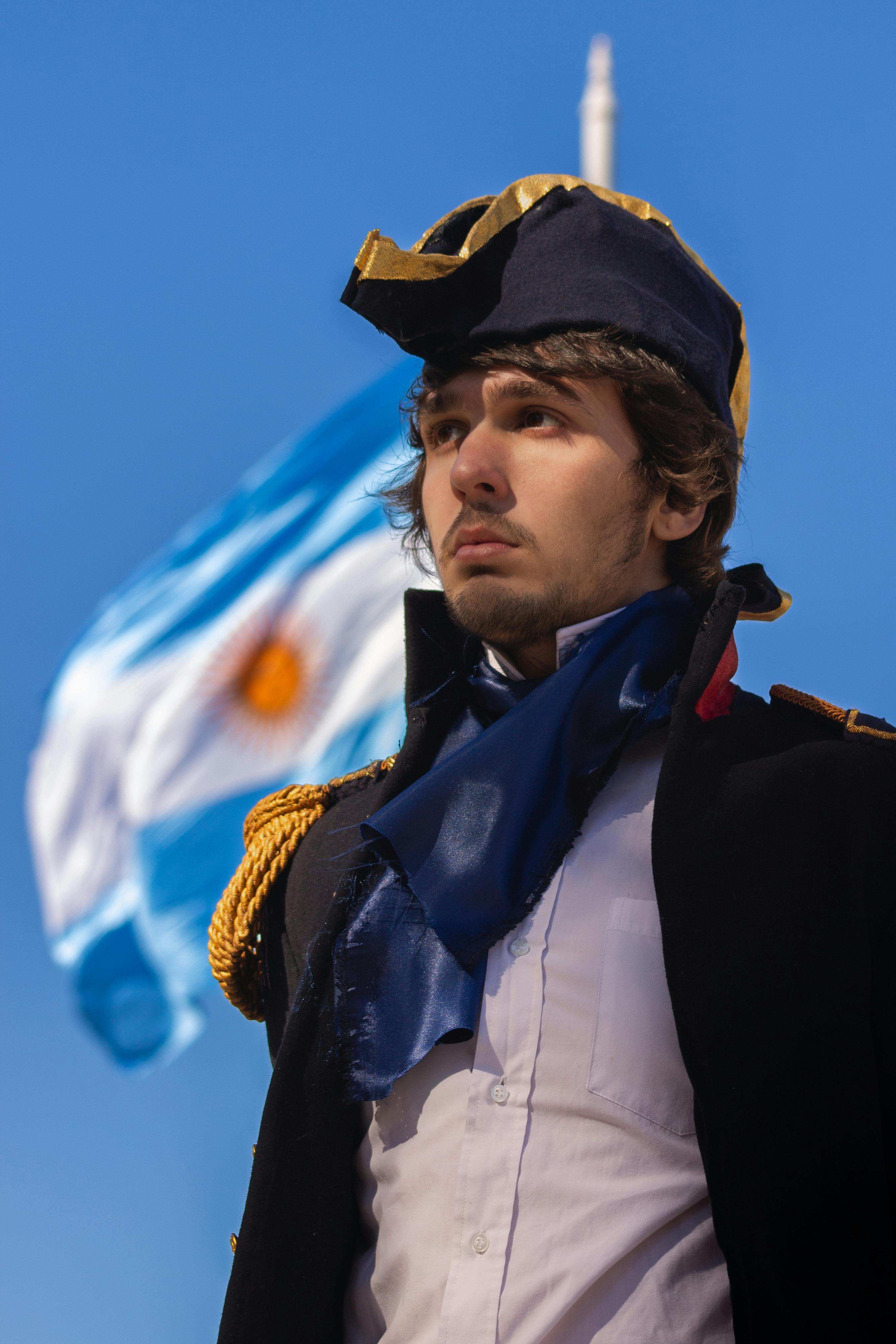 Portrait of a man in a historical costume with the Argentine flag waving in the background.