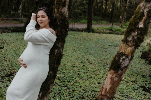 A pregnant woman in a white dress relaxes in a lush outdoor setting, leaning against a tree.