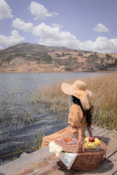 Woman in hat enjoying a lakeside picnic with a scenic mountain view. Ideal for summer lifestyle themes.