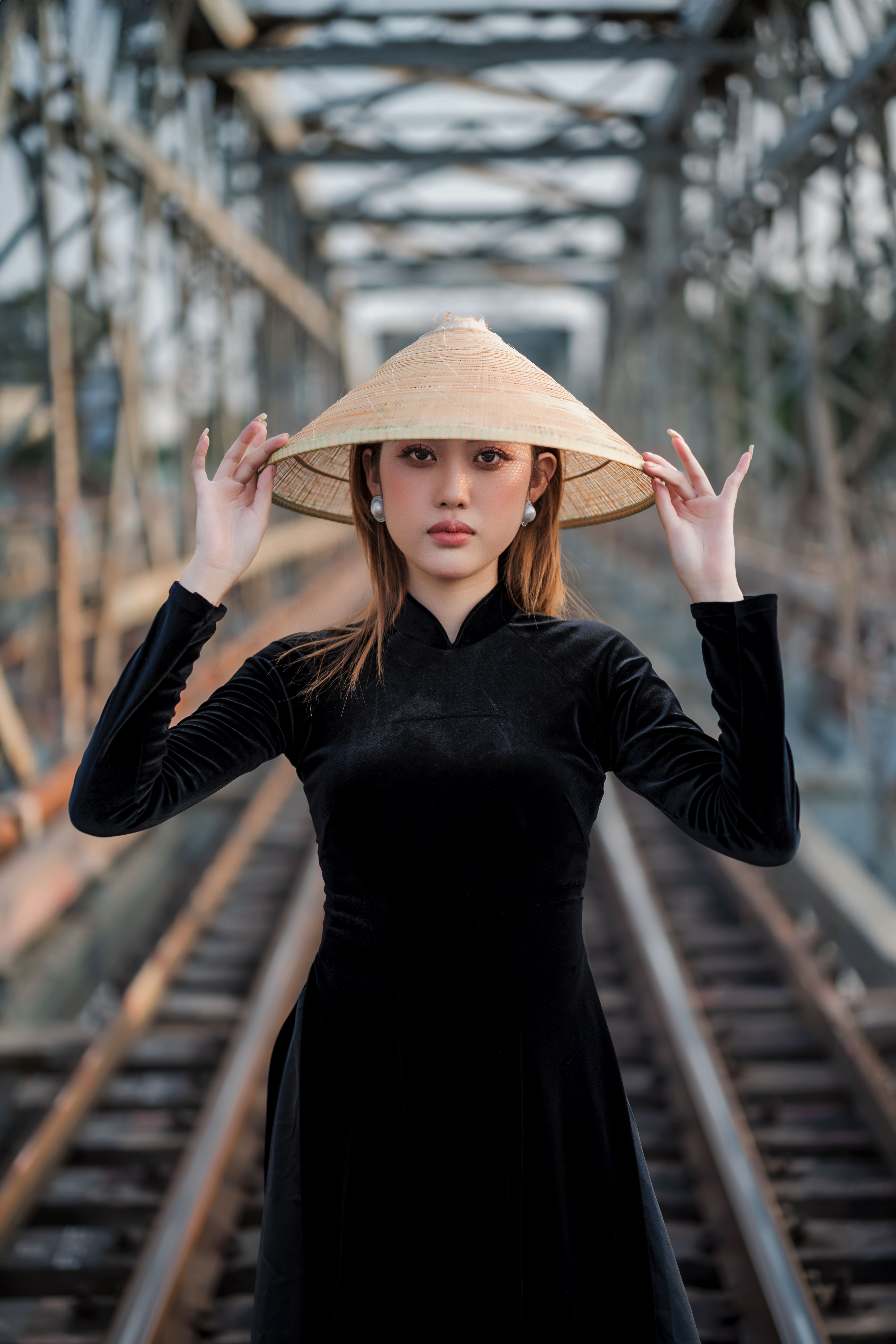 Elegant woman in ao dai holding conical hat on railway bridge, serene expression.