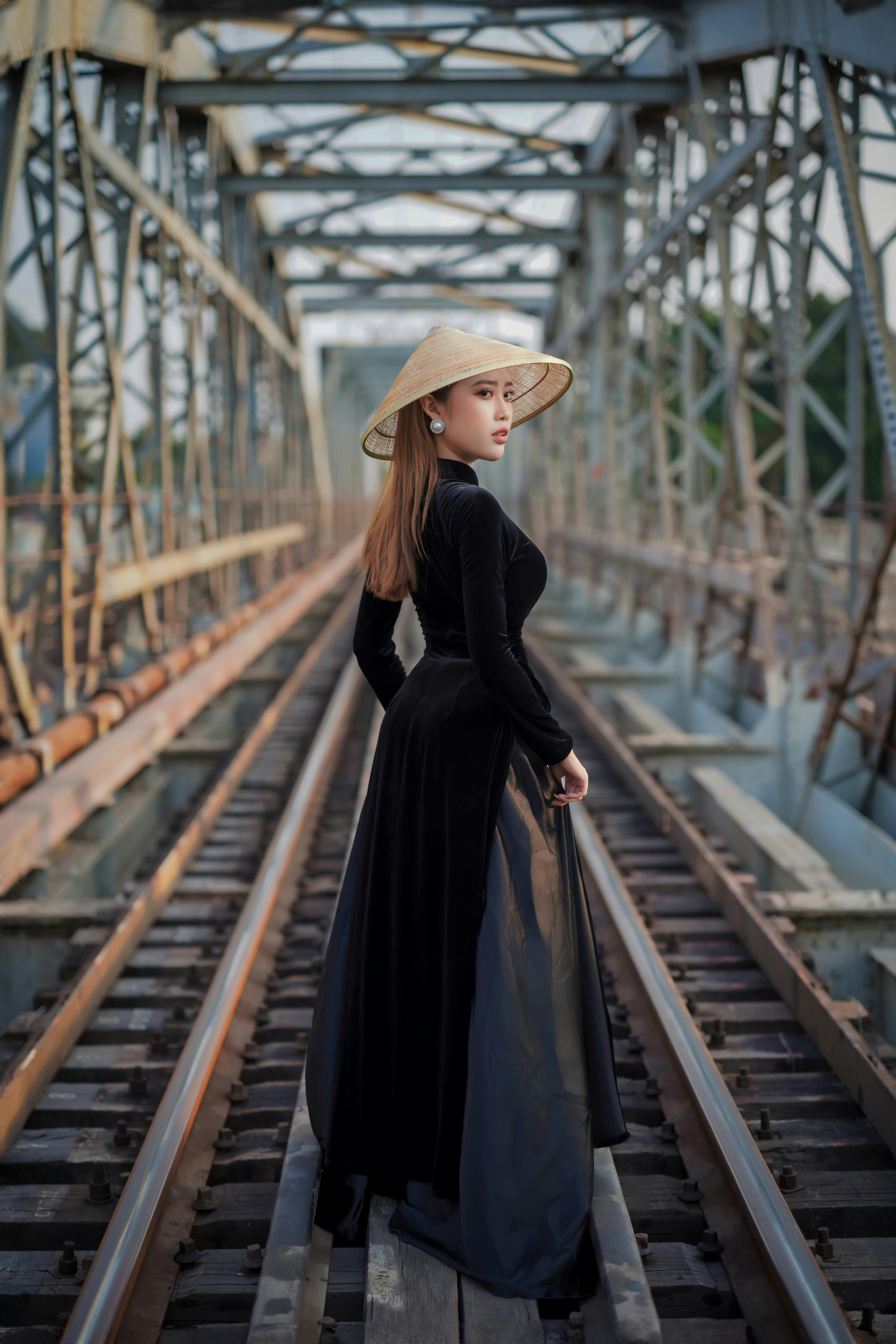 gratis Een stijlvolle vrouw in een zwarte jurk en een traditionele hoed staat op een spoorbrug en kijkt haar met serene blik aan. Stockfoto
